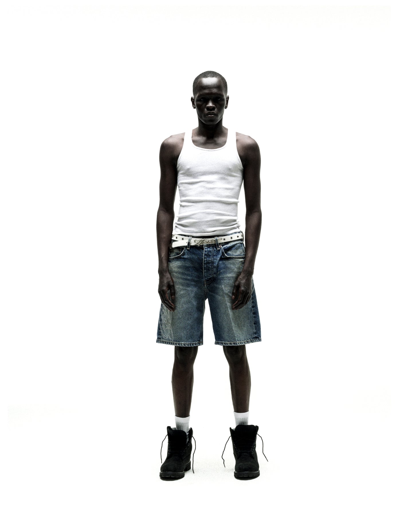 Man wearing vintage wash denim shorts with rhinestone web design, a white tank top, and black boots.