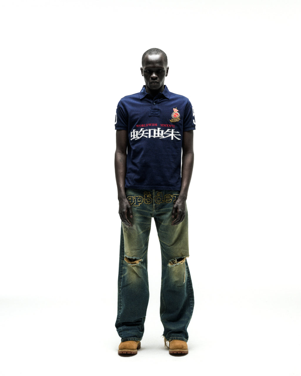 Dark-skinned model wearing a navy blue "WORLDWIDE POLO" shirt with white text and a crest, paired with distressed, wide-leg jeans and tan boots.