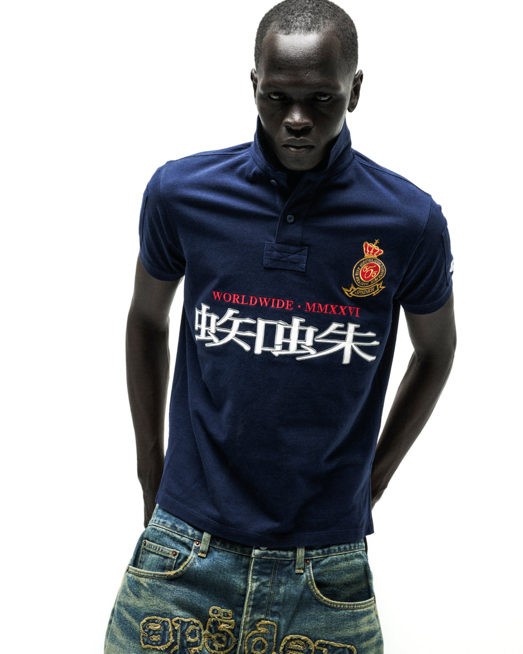 Man wearing a navy blue polo shirt with "WORLDWIDE MMXXVI" and white Chinese characters, paired with distressed jeans.