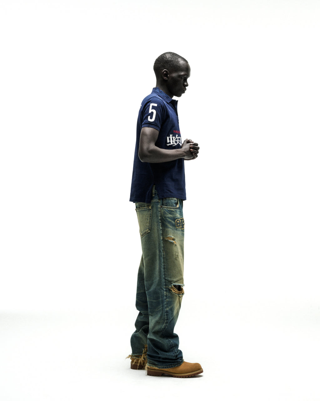 Man in navy WORLDWIDE POLO shirt with white "5" and red Japanese text, paired with distressed blue jeans and brown boots.