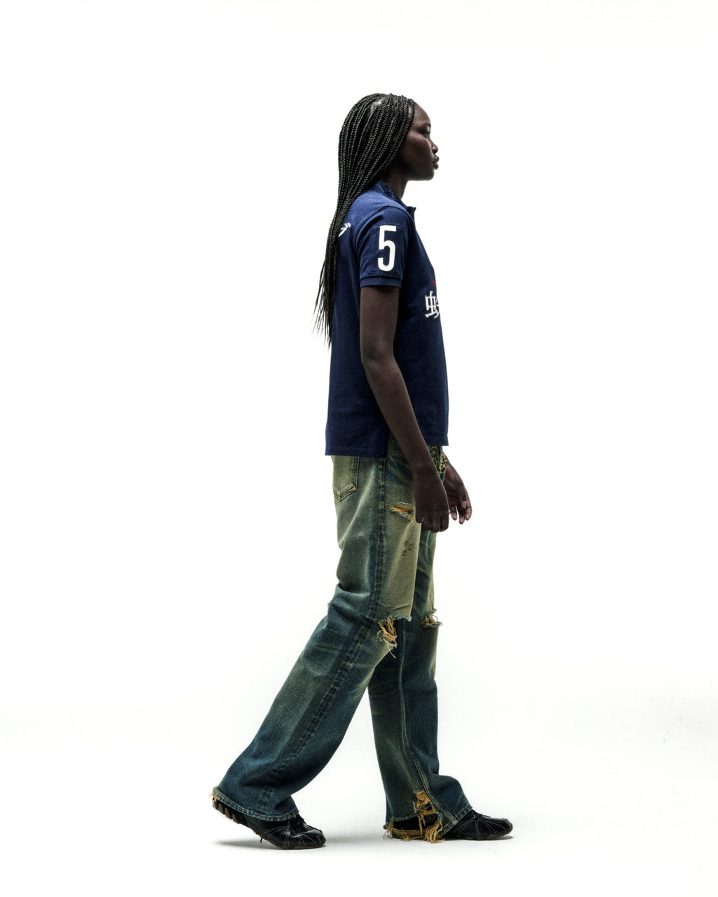 Dark-skinned model in a navy blue polo shirt with "5" and white text, paired with distressed denim jeans.