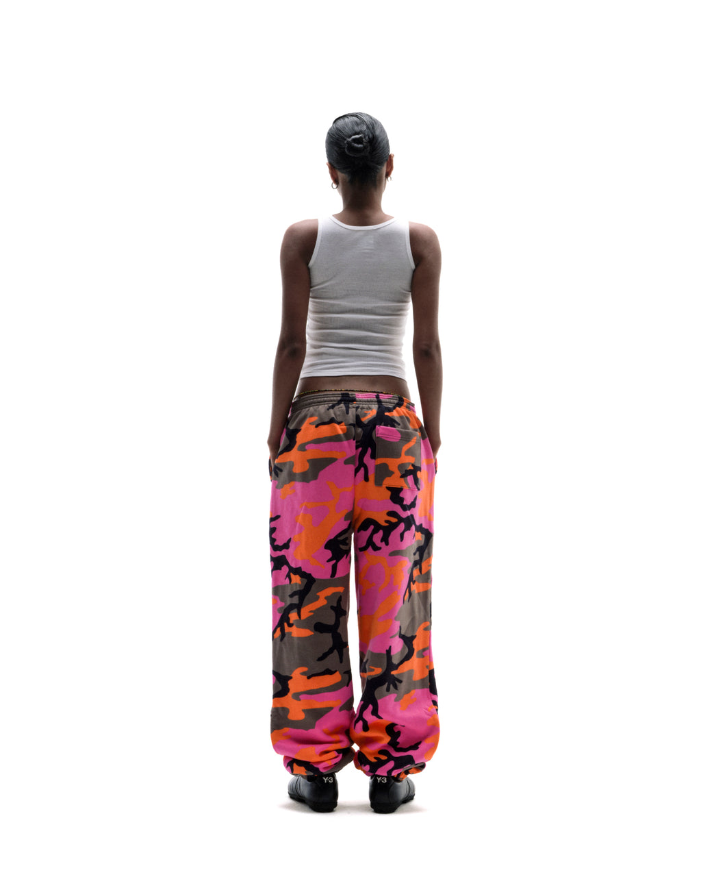 Model seen from behind wearing a white ribbed tank top and vibrant pink and orange camouflage sweatpants.