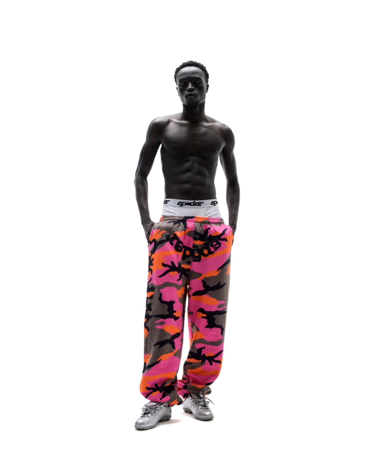 Model wearing pink and orange camouflage sweatpants with a white branded waistband and silver sneakers against white.