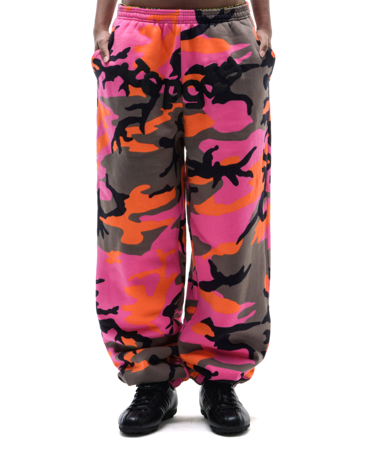 Model wearing loose-fitting fleece sweatpants in a vibrant pink, orange, and grey camouflage print with black sneakers.