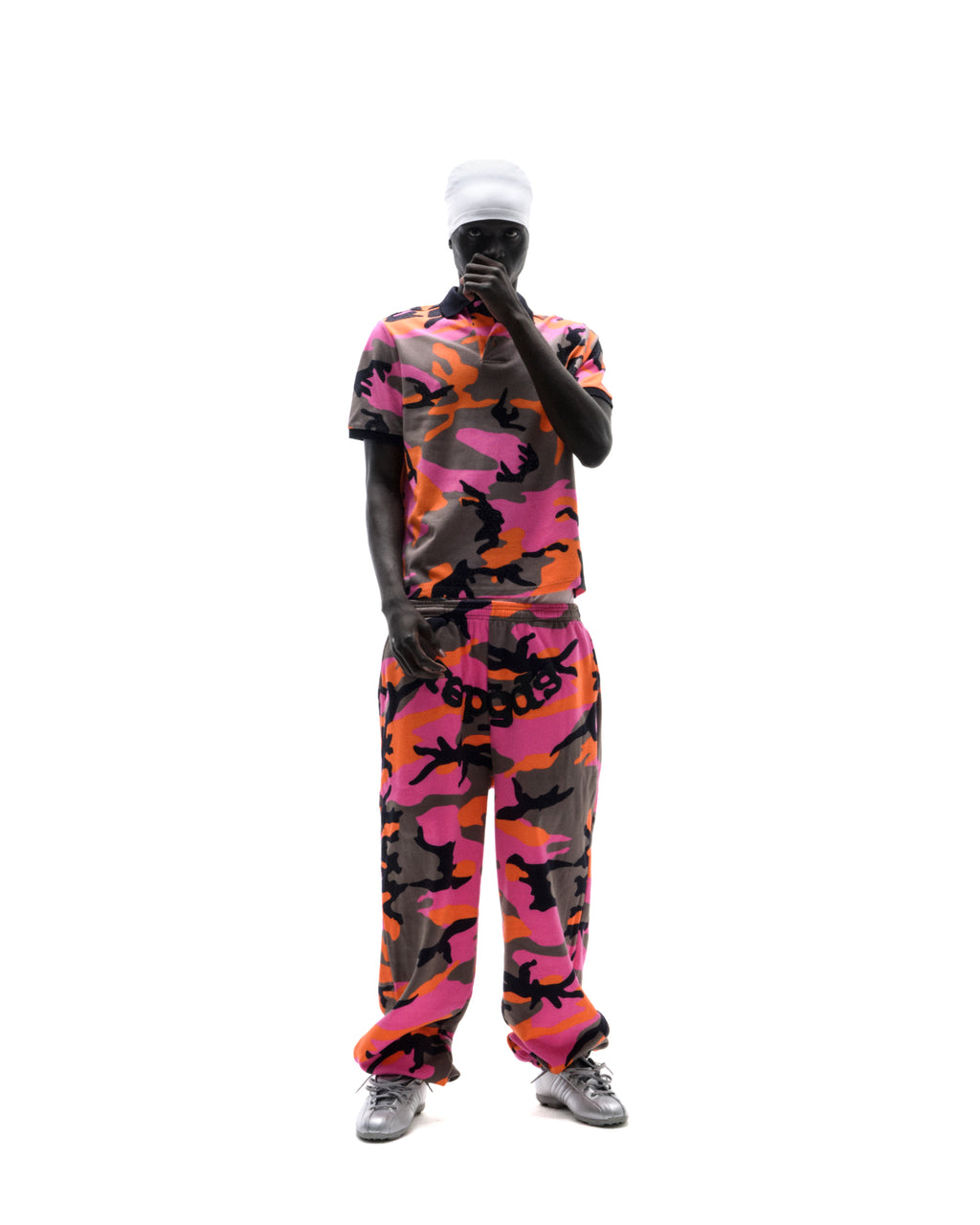 Model wearing a pink, orange, and grey camouflage polo shirt and matching sweatpants with a white beanie and silver sneakers.