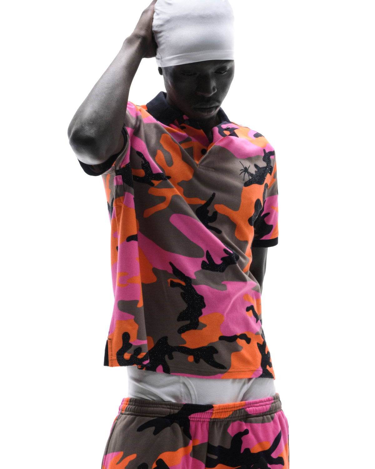 Model wearing a pink, orange, and brown camouflage polo shirt and matching shorts with a white head covering.