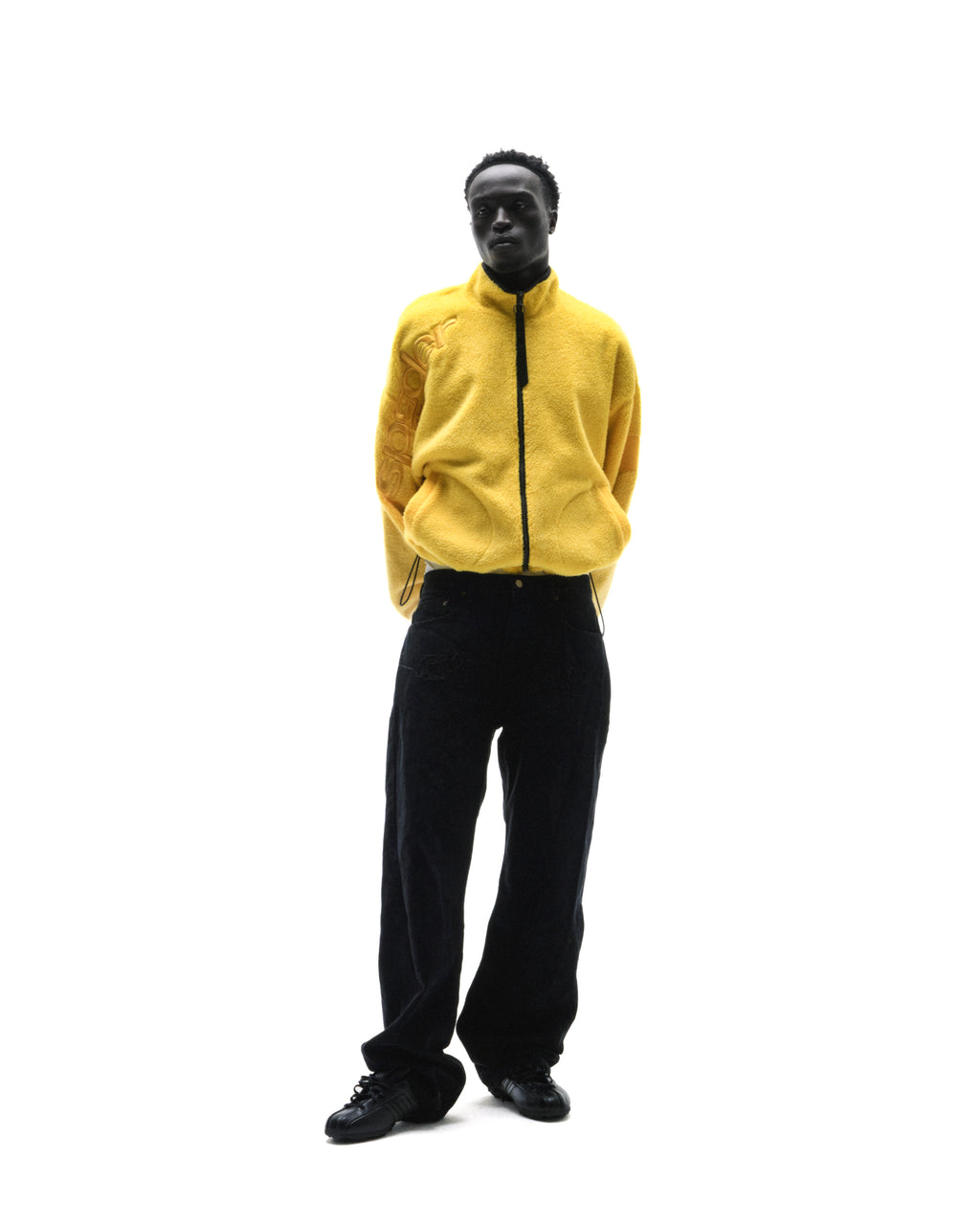 A model wearing a bright yellow zip-up fleece jacket and black trousers, standing against a plain white background.