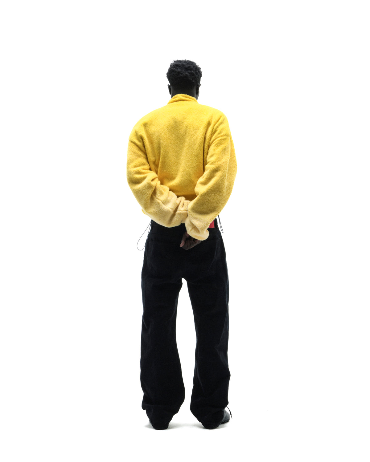A model seen from behind wearing a bright yellow fleece sweater and loose-fitting black trousers against a white background.
