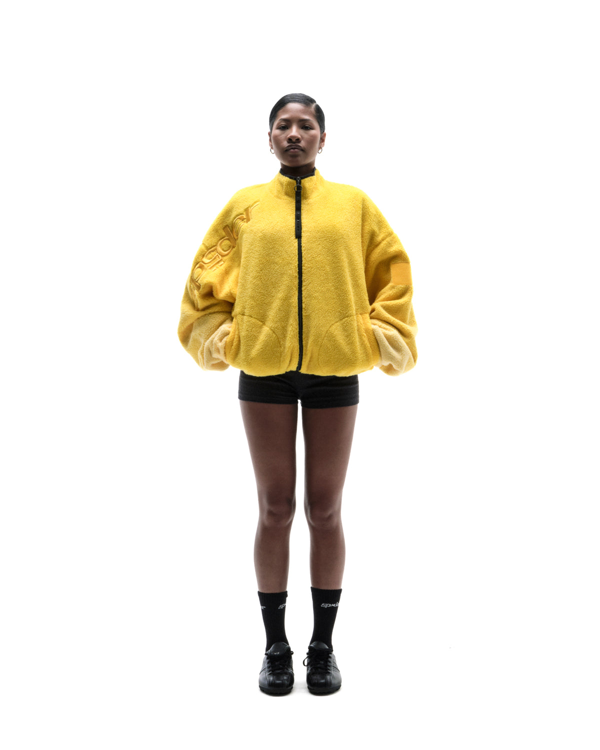 Model wearing a bright yellow oversized fleece jacket with a black zipper, black shorts, and black sneakers.
