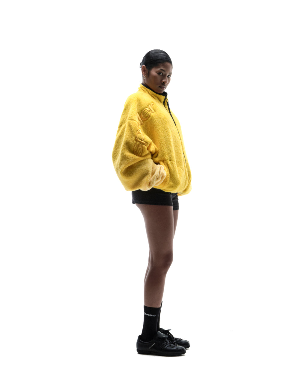 Model wearing a bright yellow oversized fleece jacket with embossed branding, black shorts, and black sneakers.