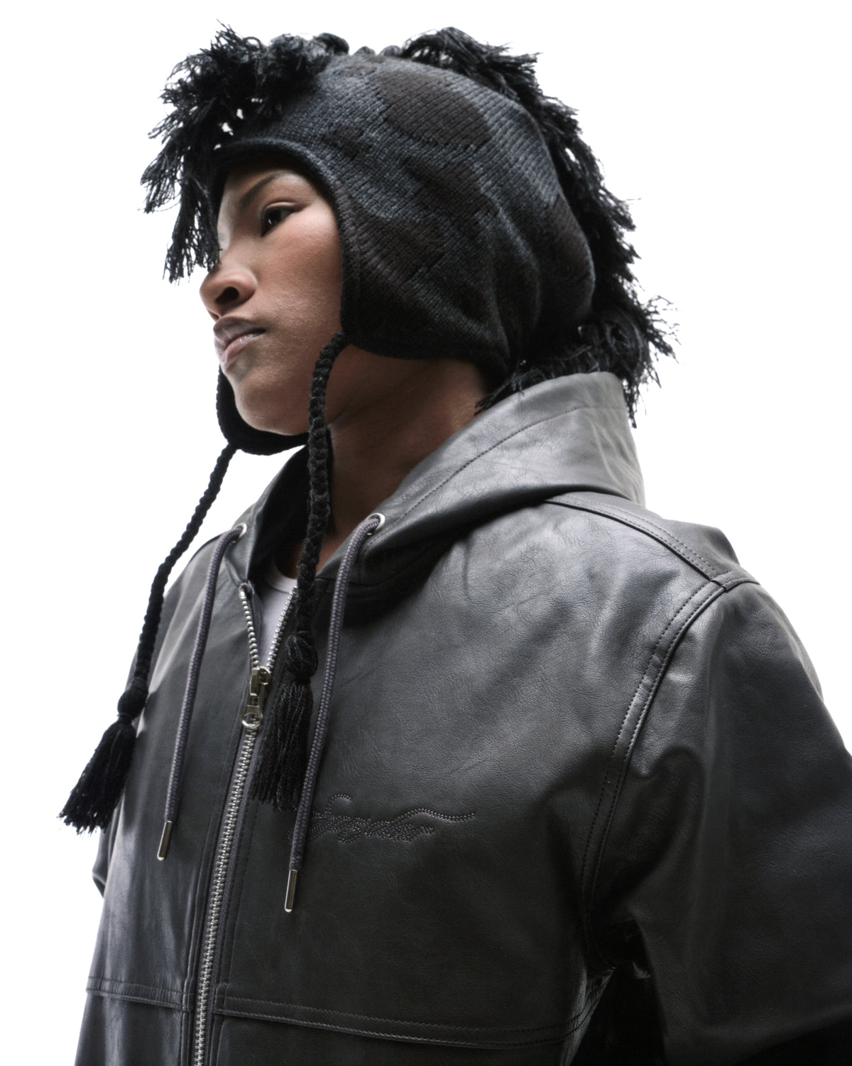 Model wearing a black leather hooded jacket and a textured black knit hat with braided tassels against a white background.