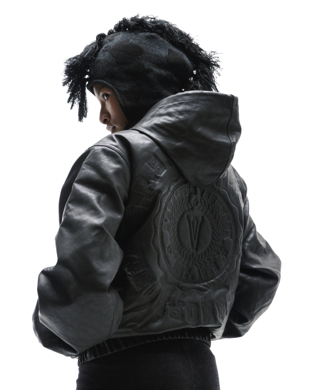 A model wearing a black leather hooded jacket with a large circular embossed logo on the back and a textured knit hat.
