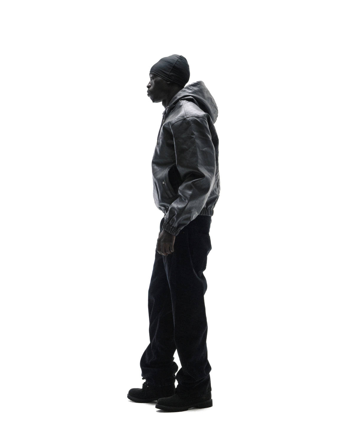 Model in profile wearing a black leather hooded jacket, dark trousers, and a beanie against a white background.