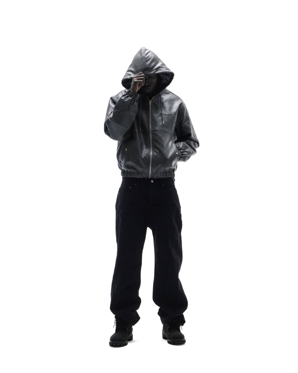 Model wearing a black leather hooded jacket and loose-fit dark trousers against a white background.