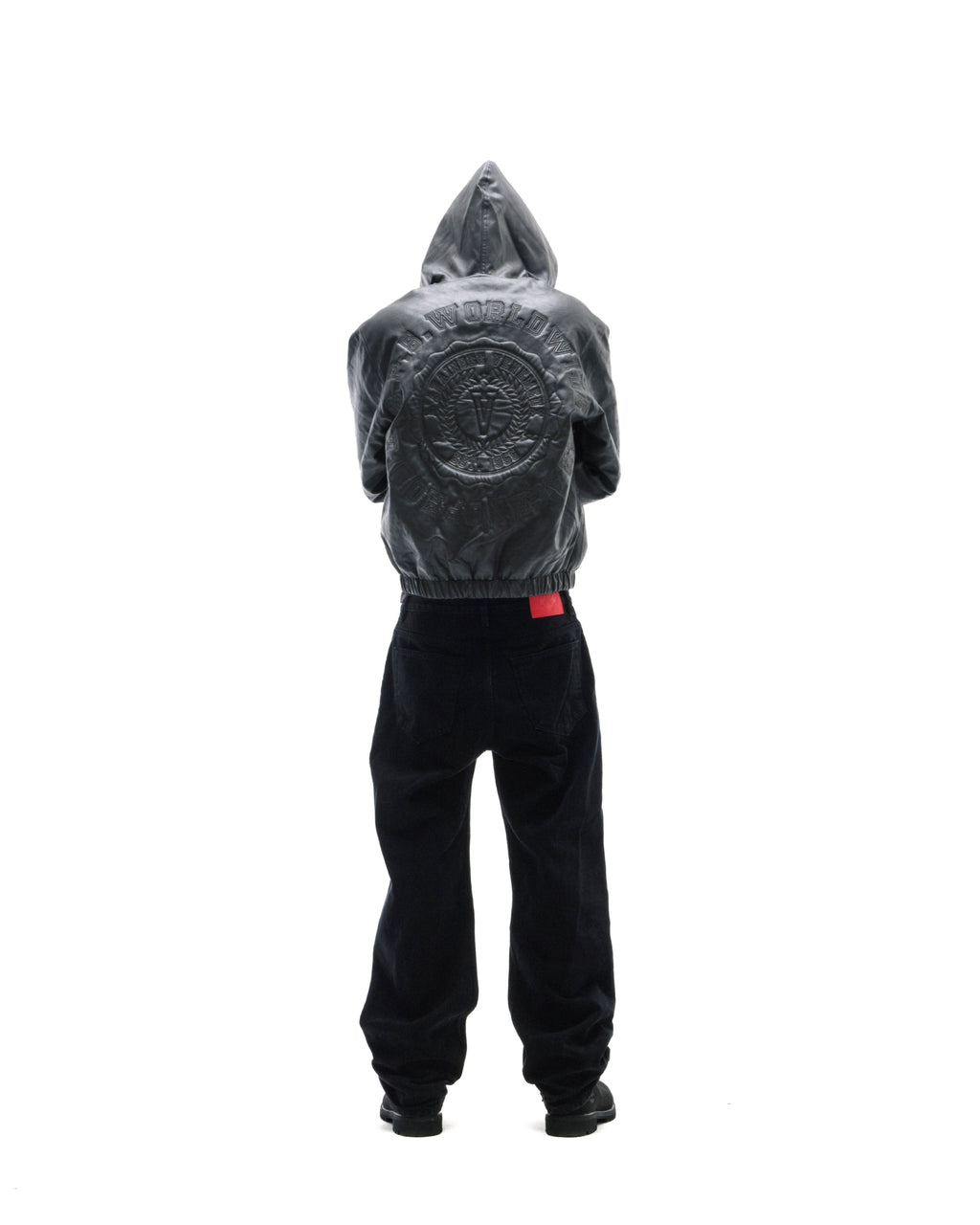 Model wearing a black leather hooded jacket with embossed back detailing and loose-fit black jeans against white.