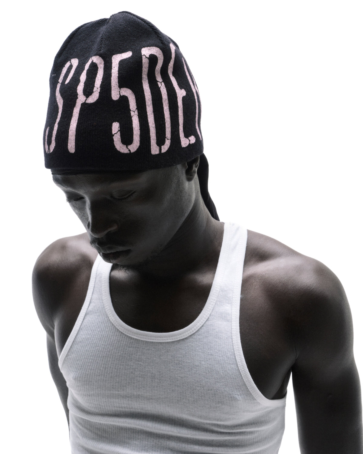 Model wearing a black beanie with pink SP5DER lettering and a white ribbed tank top against a white background.