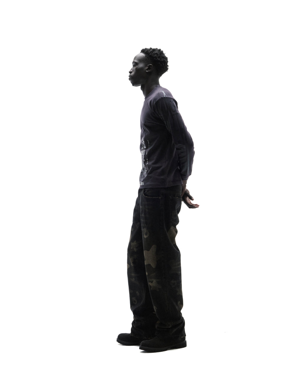Model in profile wearing a dark long-sleeve graphic t-shirt, loose-fit camouflage print jeans, and black boots.