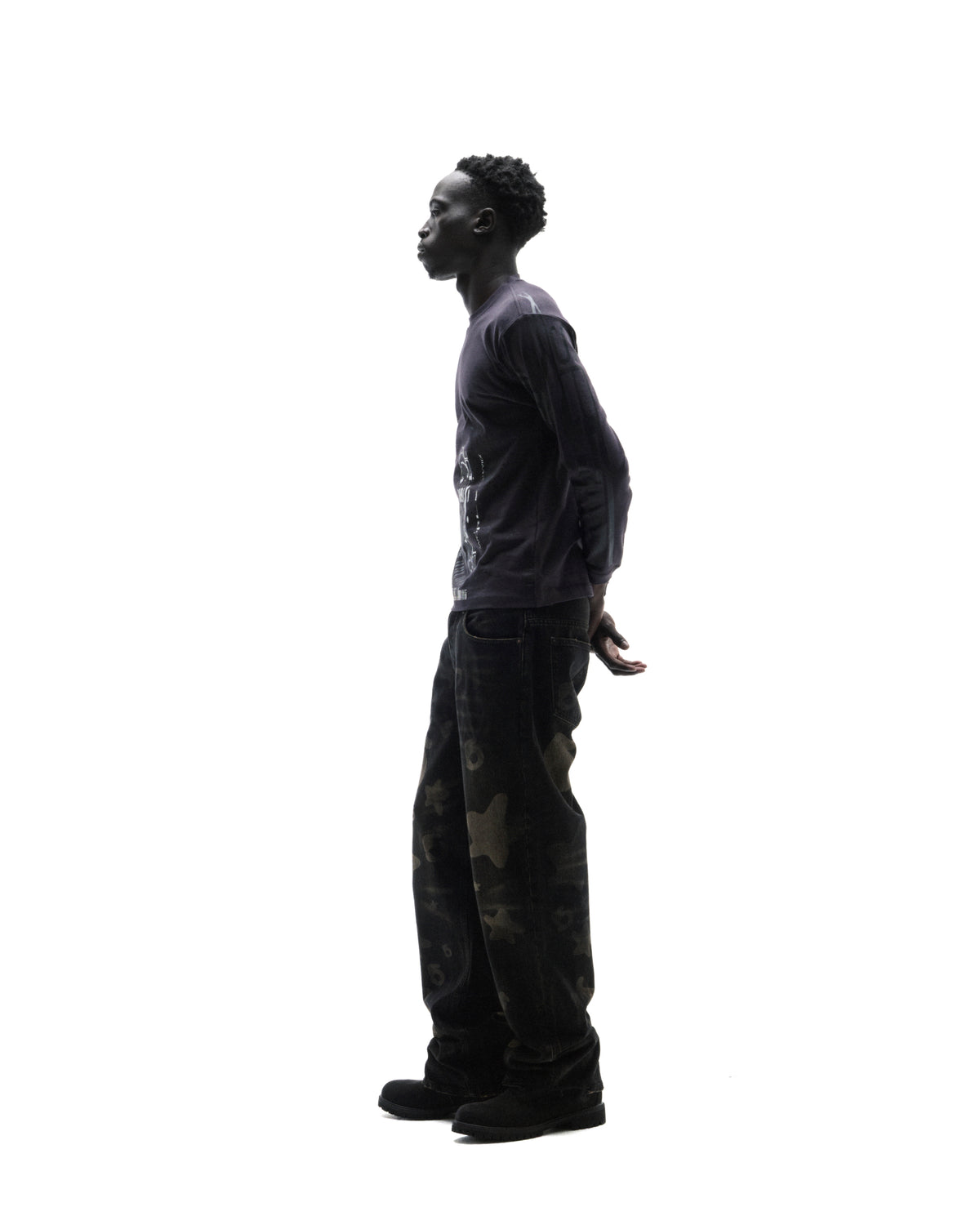 Model in profile wearing a dark long-sleeve graphic t-shirt, loose-fit camouflage print jeans, and black boots.