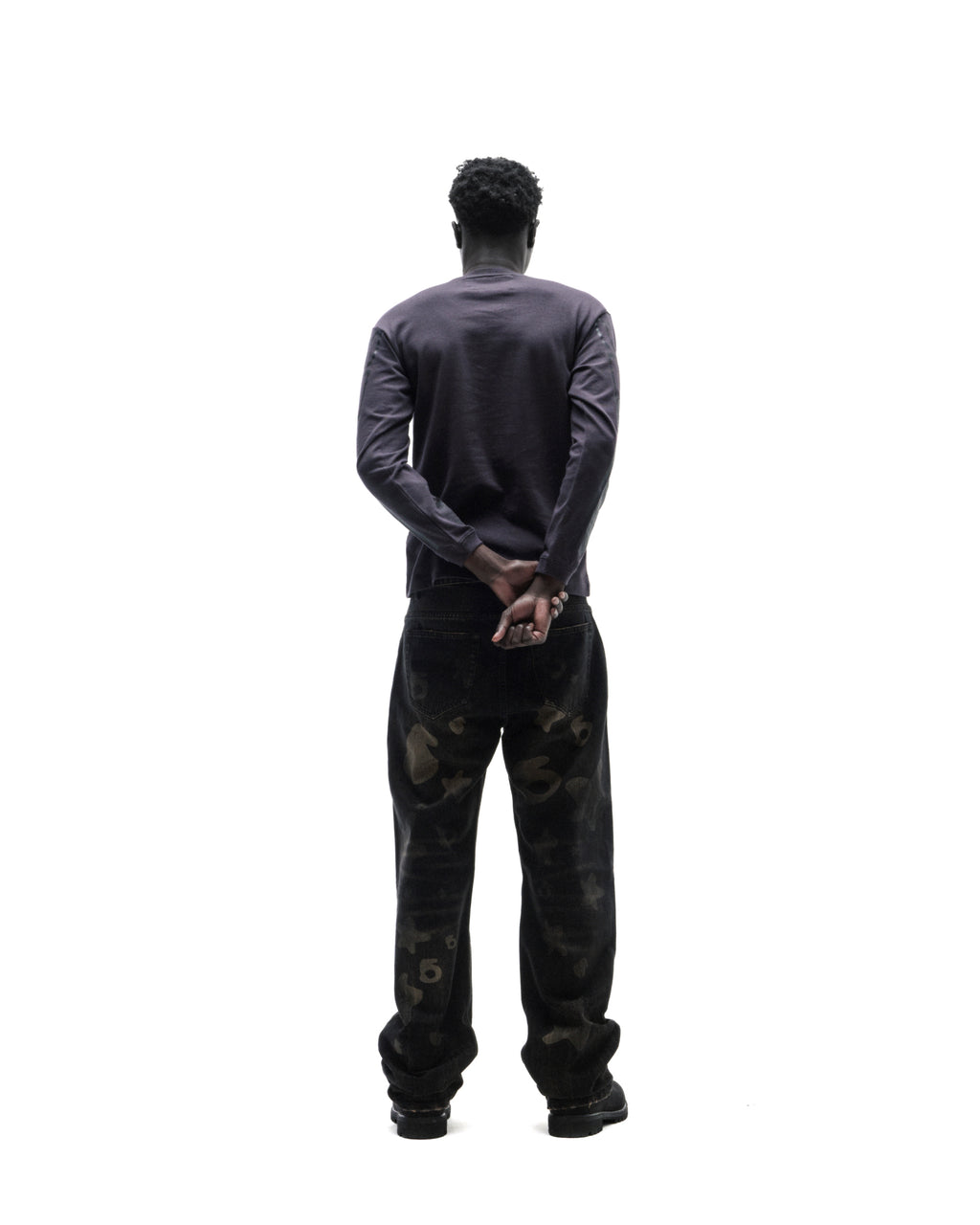 Model seen from behind wearing a dark long-sleeve shirt and black patterned denim jeans against a white background.