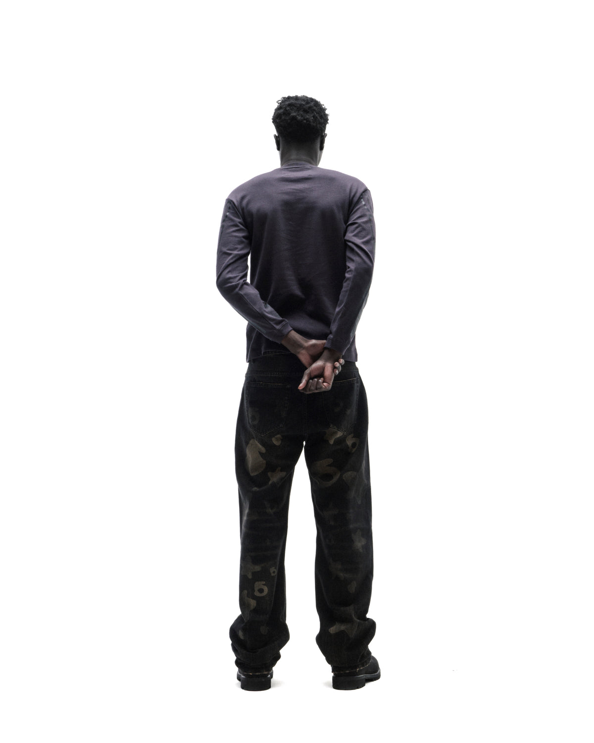 Model seen from behind wearing a dark long-sleeve shirt and black patterned denim jeans against a white background.