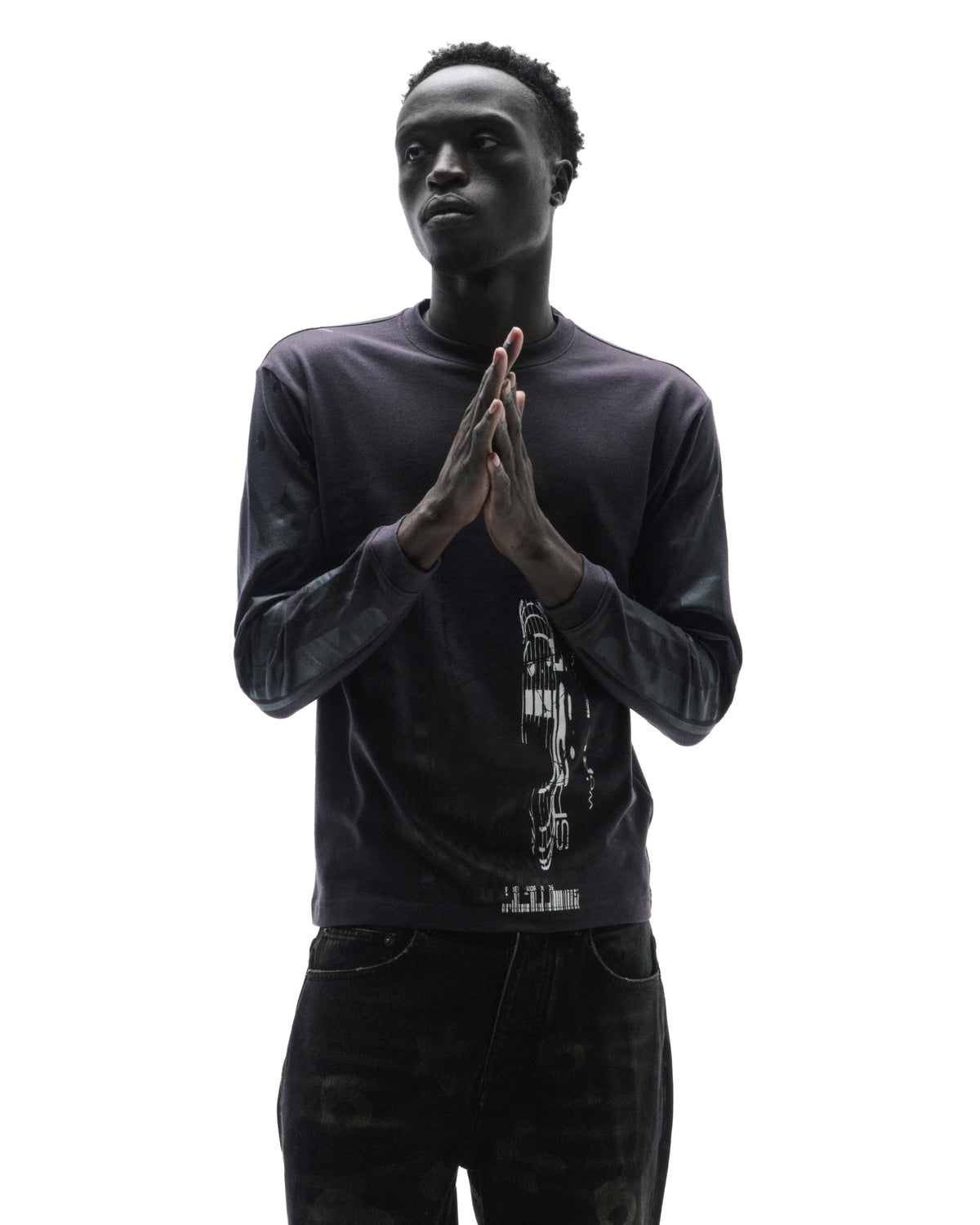 A model wearing a dark long-sleeve graphic t-shirt and black jeans, posing with hands pressed together against a white background.