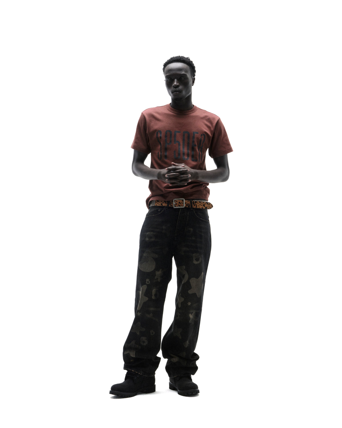 Model wearing a brown graphic t-shirt, patterned black baggy jeans, and a leopard print belt against a white background.