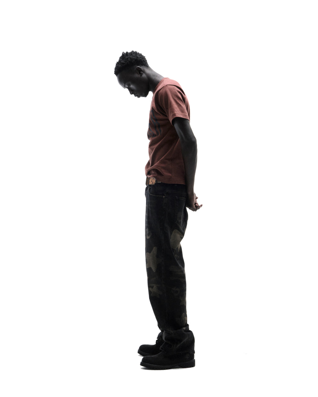 A model stands in profile wearing a brown t-shirt, patterned black jeans, and black boots against a white background.