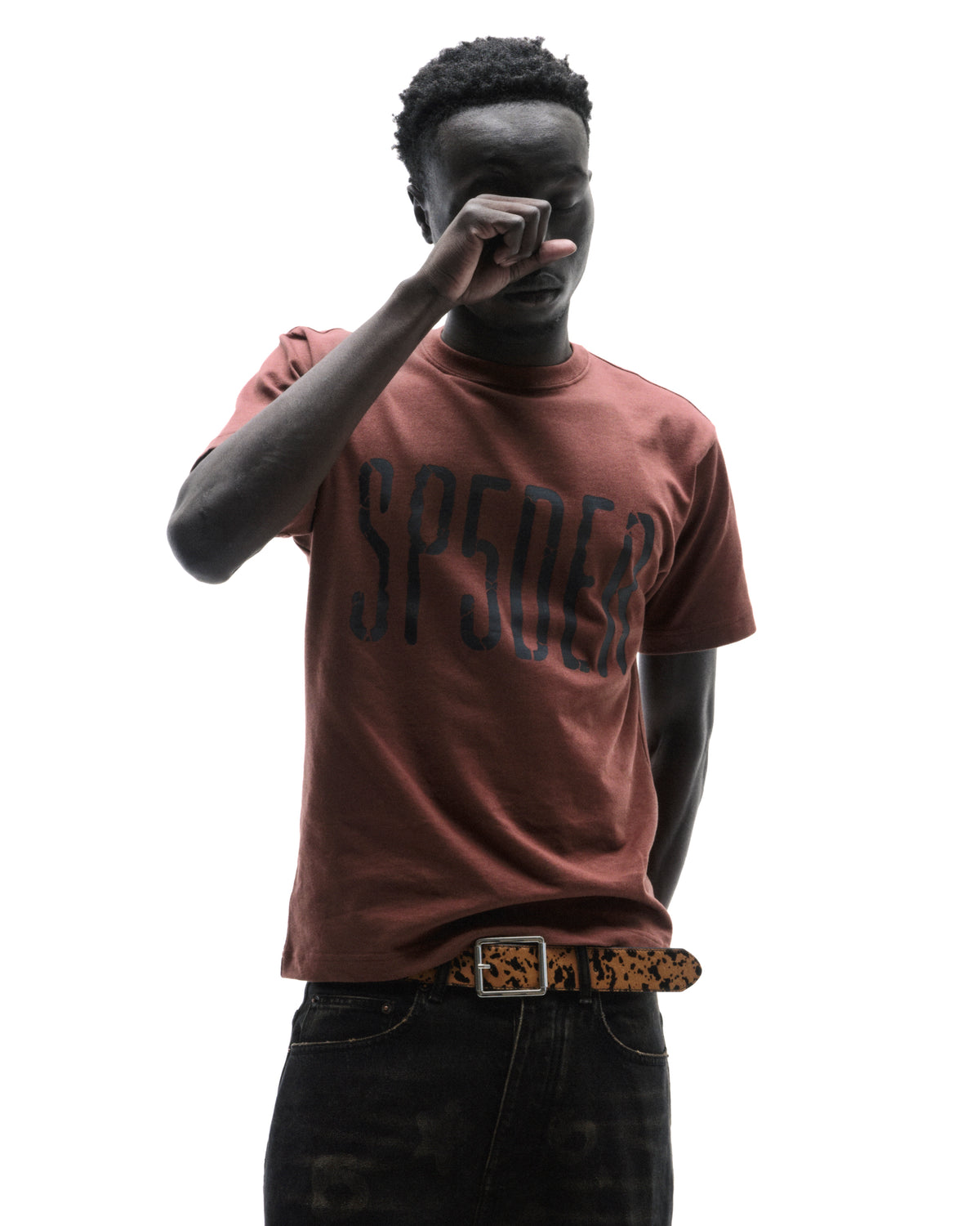 A model wearing a brown SP5DER graphic t-shirt, black jeans, and a leopard print belt against a white background.