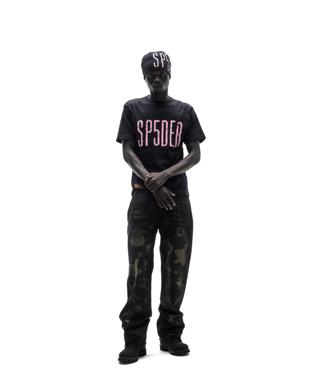 Model wearing a black SP5DER t-shirt, matching beanie, and dark patterned jeans against a white background.
