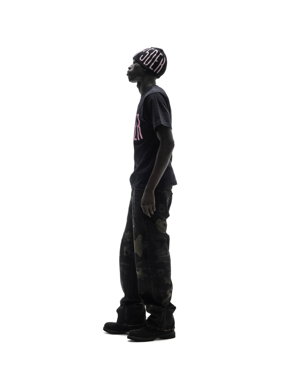 Model in a black beanie and t-shirt with pink lettering, wearing dark camo-patterned baggy jeans against a white background.