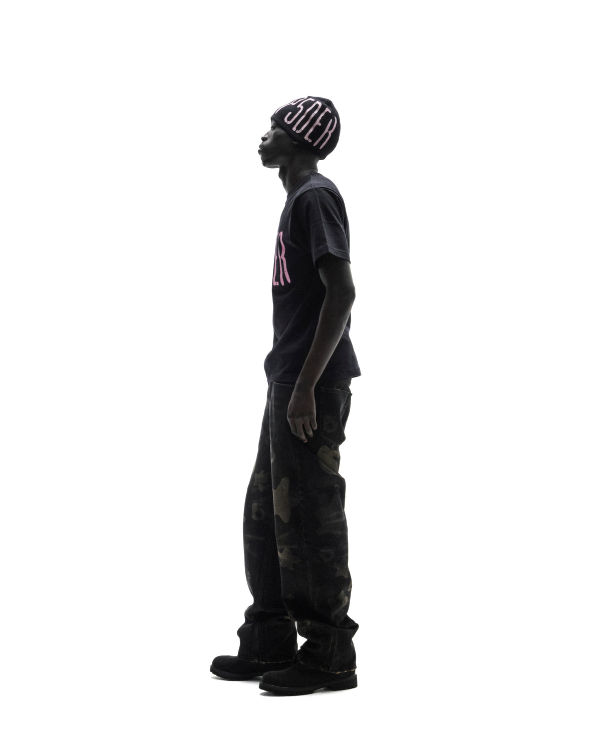 Model in a black beanie and t-shirt with pink lettering, wearing dark camo-patterned baggy jeans against a white background.