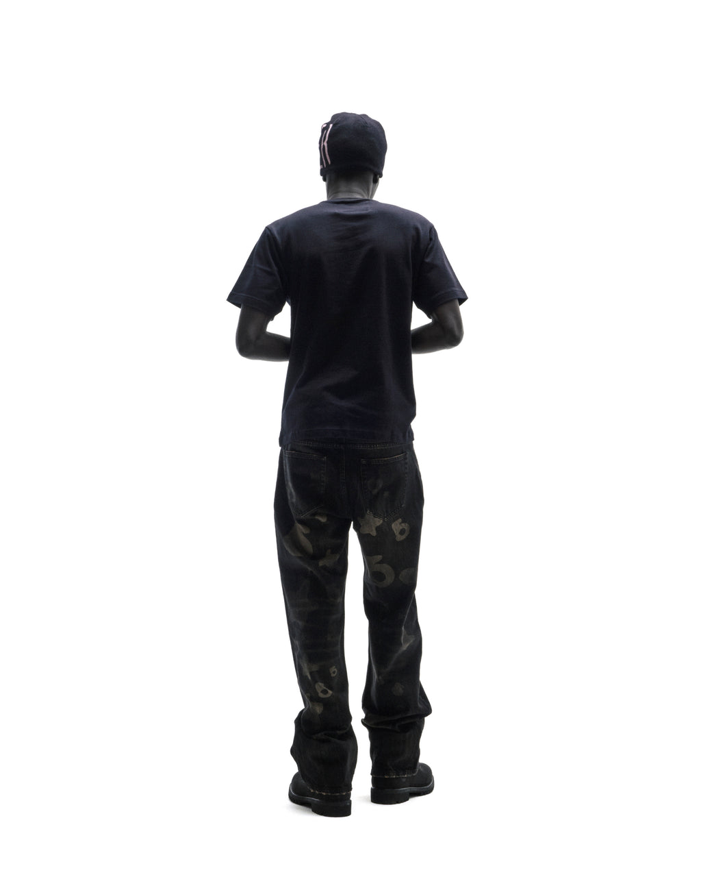 A model seen from behind wearing a black t-shirt, dark patterned jeans, and a black beanie against a white background.