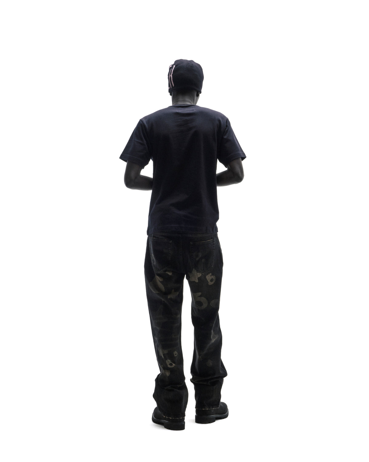 A model seen from behind wearing a black t-shirt, dark patterned jeans, and a black beanie against a white background.
