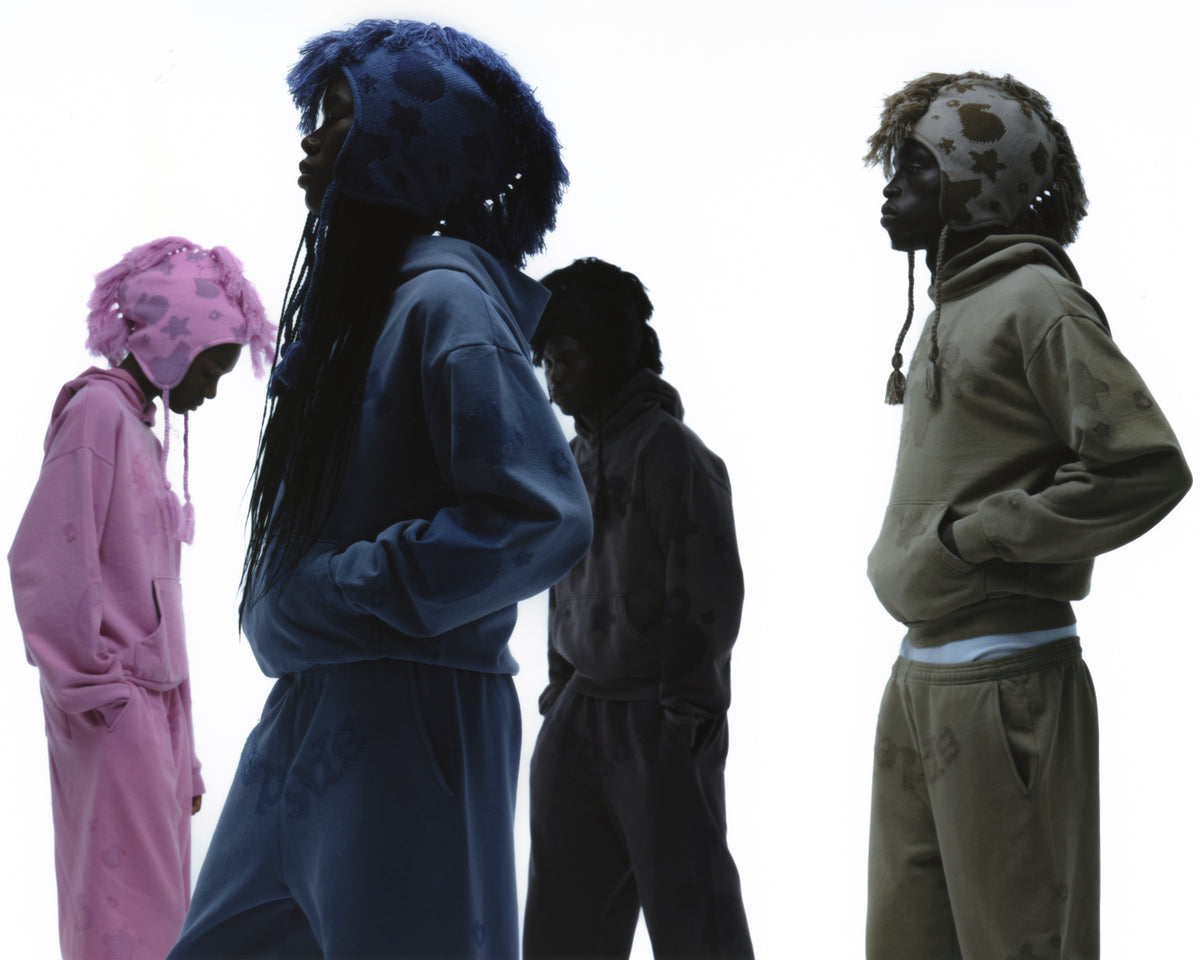Four models wearing matching hooded tracksuits in different muted colors—pink, blue, black, and olive green—standing in profile against a solid white background.

