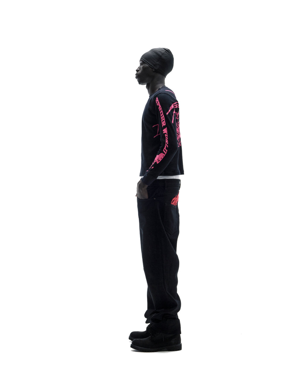 Model wearing a black long-sleeve shirt with pink graphics, black jeans, and a beanie against a white background.