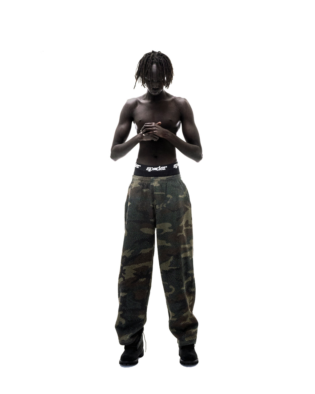 Model with dreadlocks wearing camouflage mesh pants and black Sp5der branded underwear against a white background.
