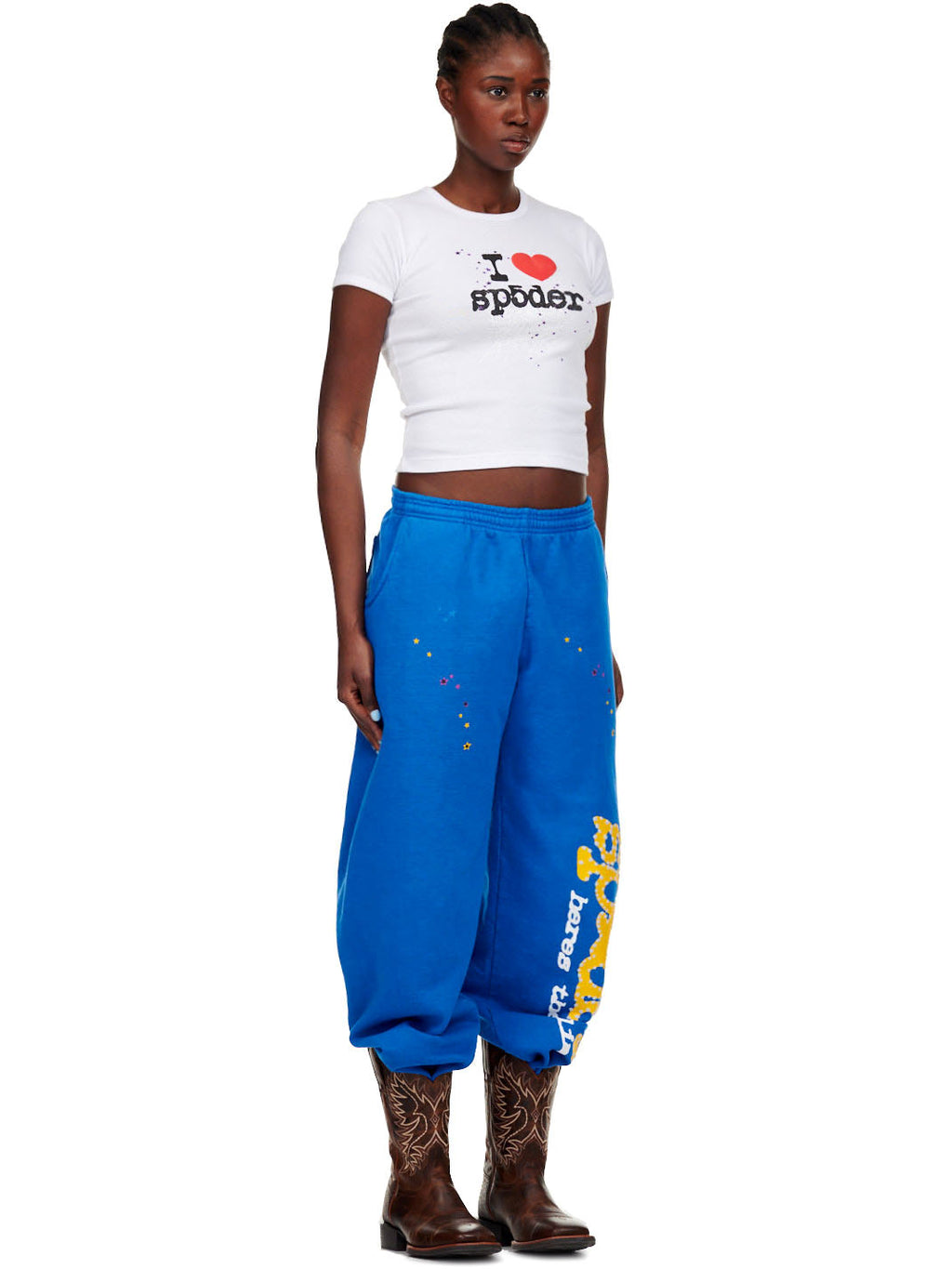 Female model in blue TC BLUE SWEATPANT with yellow SP5DER! text and star print, paired with a white tee and cowboy boots.