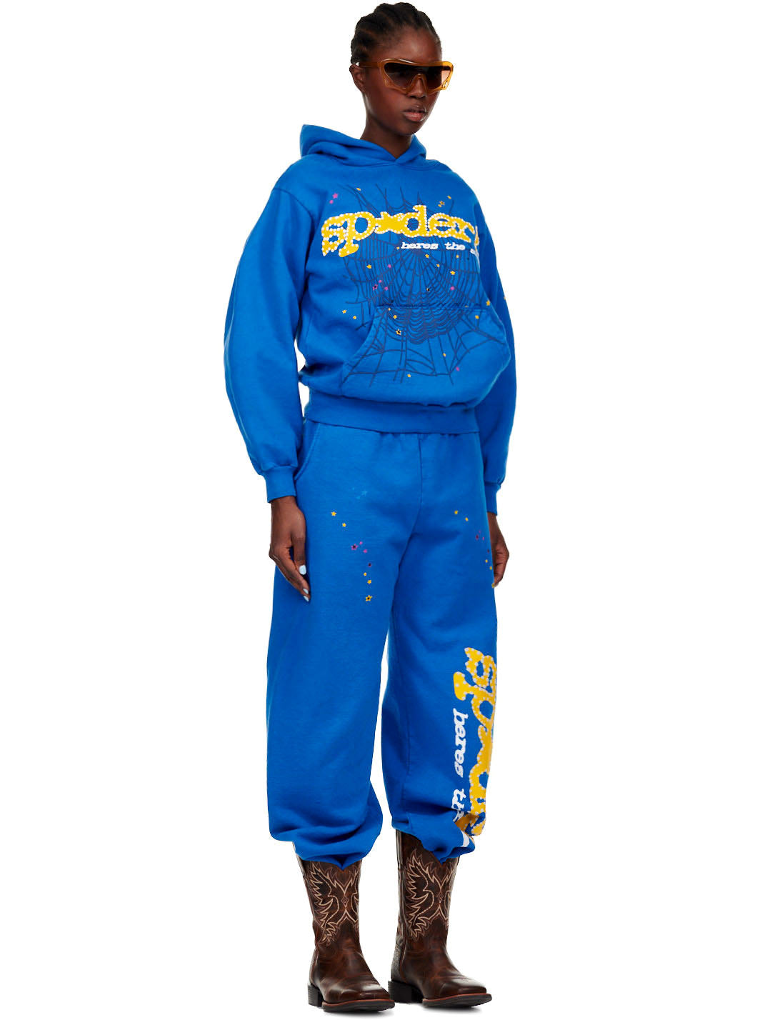 Female model in blue fleece hoodie and sweatpants with SP5DER! text, web, and star prints.