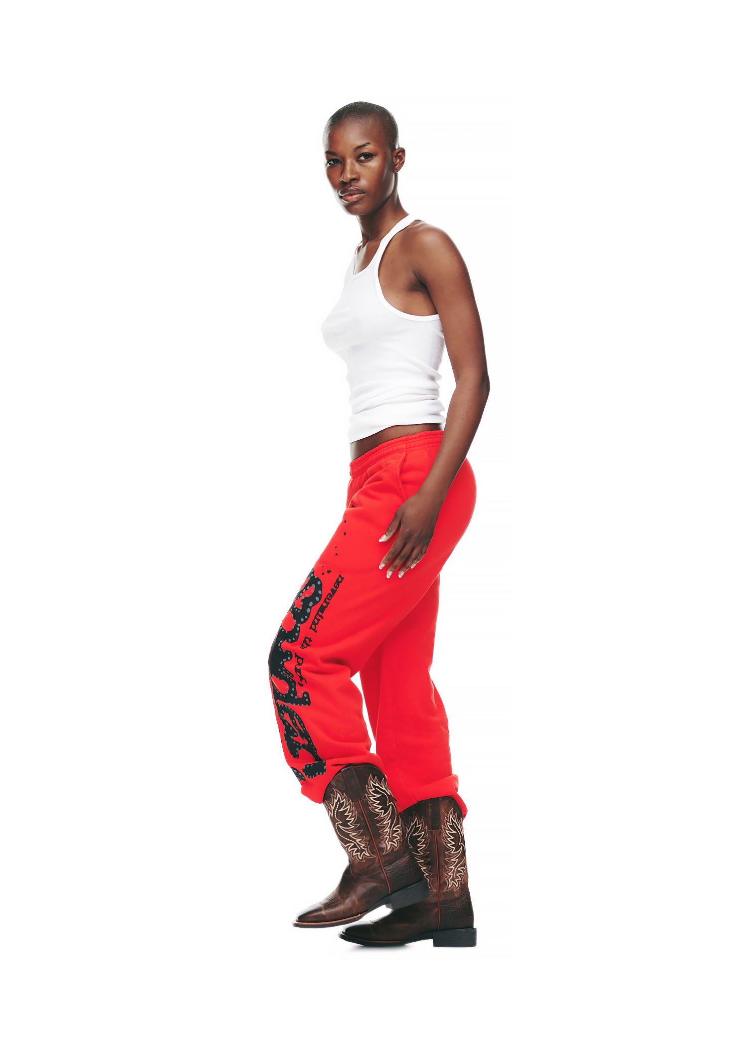 Female model in red P*NK V2 sweatpants with black SP5DER puff print and star motifs, white tank top, and cowboy boots.