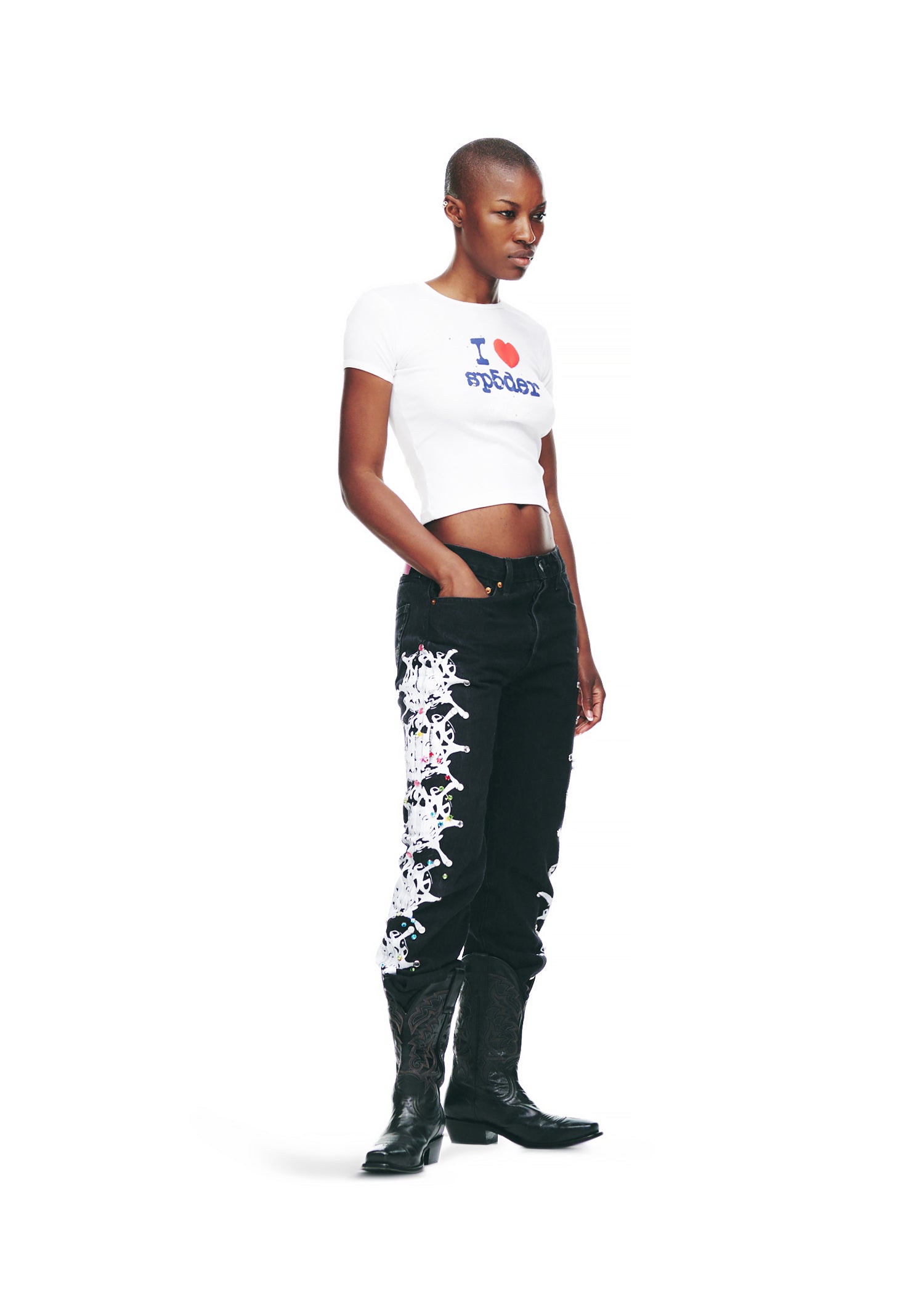 Female model in white cropped cotton baby tee with "I ❤️ Sp5der" graphic, black jeans, and cowboy boots.