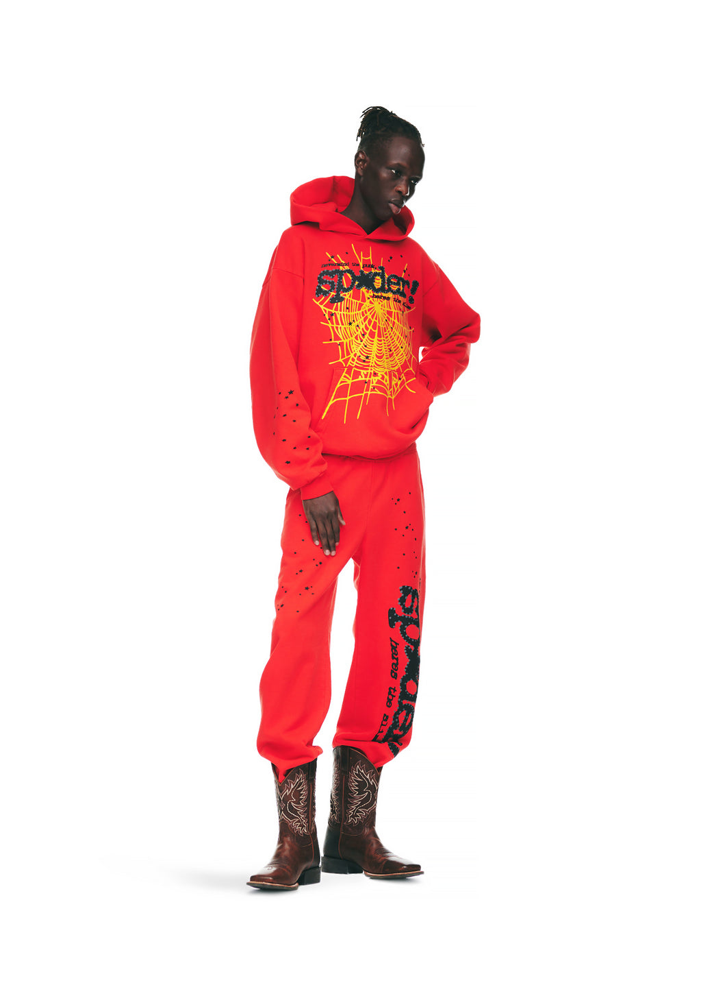 Male model in red P*NK V2 hoodie and sweatpants with yellow spiderweb and black SP5DER puff print, wearing cowboy boots.