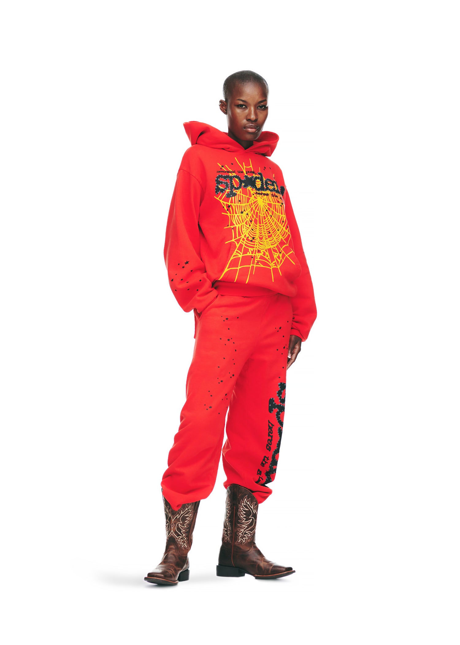 Female model in a red P*NK V2 hoodie and sweatpants with yellow spider web and SP5DER prints.