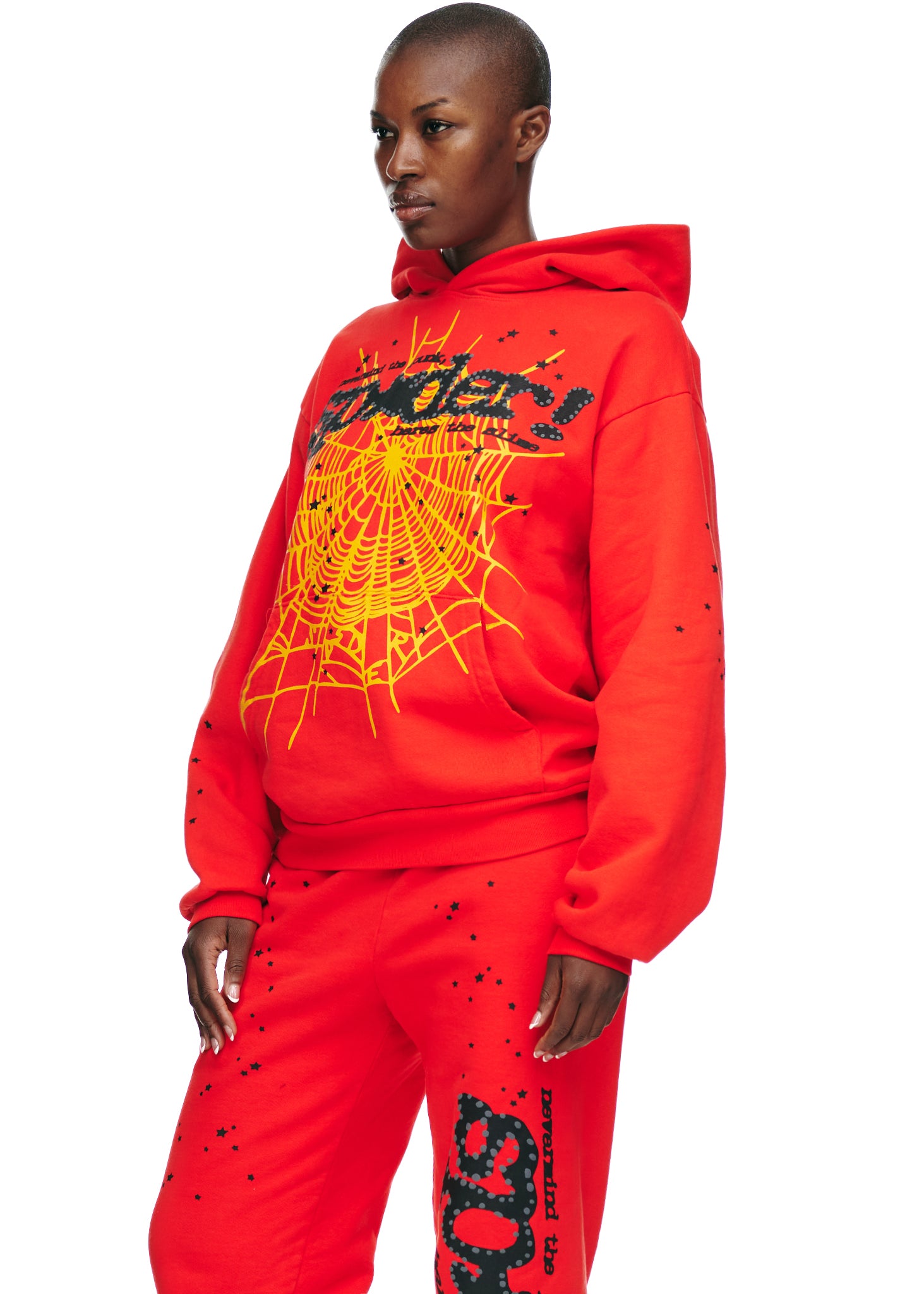 Female model in red P*NK V2 HOODIE with yellow spider web, SP5DER motif, and star prints.