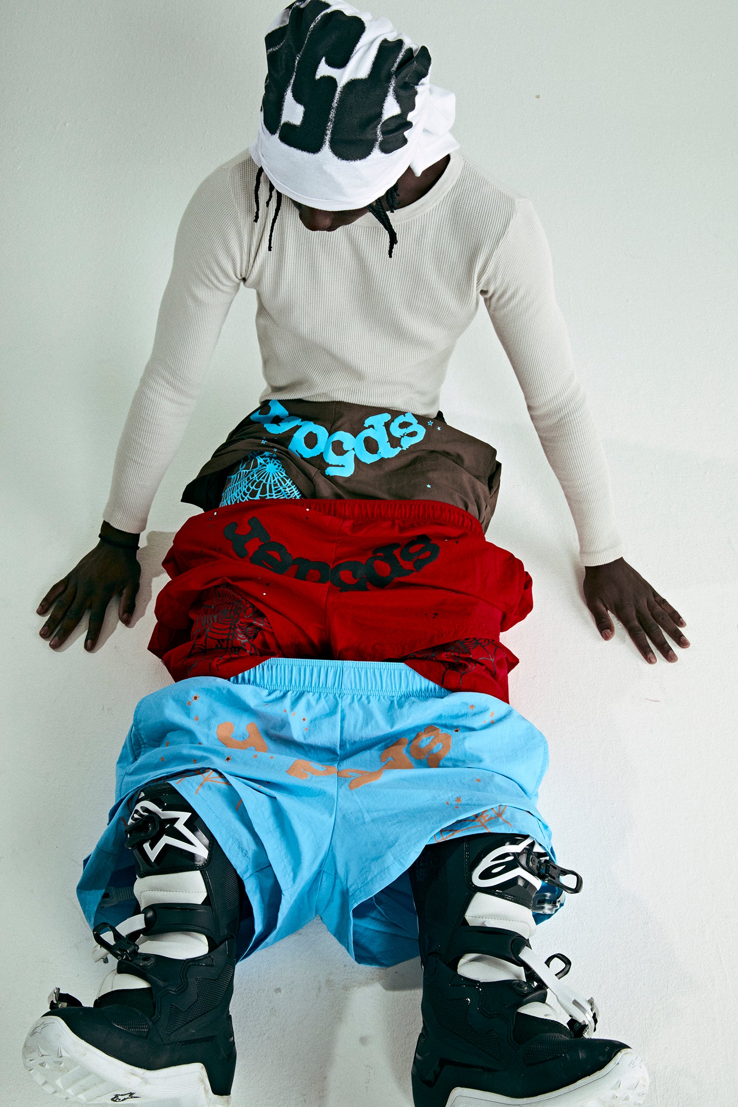 Model wearing a white long-sleeve shirt, white beanie, and stacked OG WEB DOUBLE LAYER SHORTS in brown, red, and blue.
