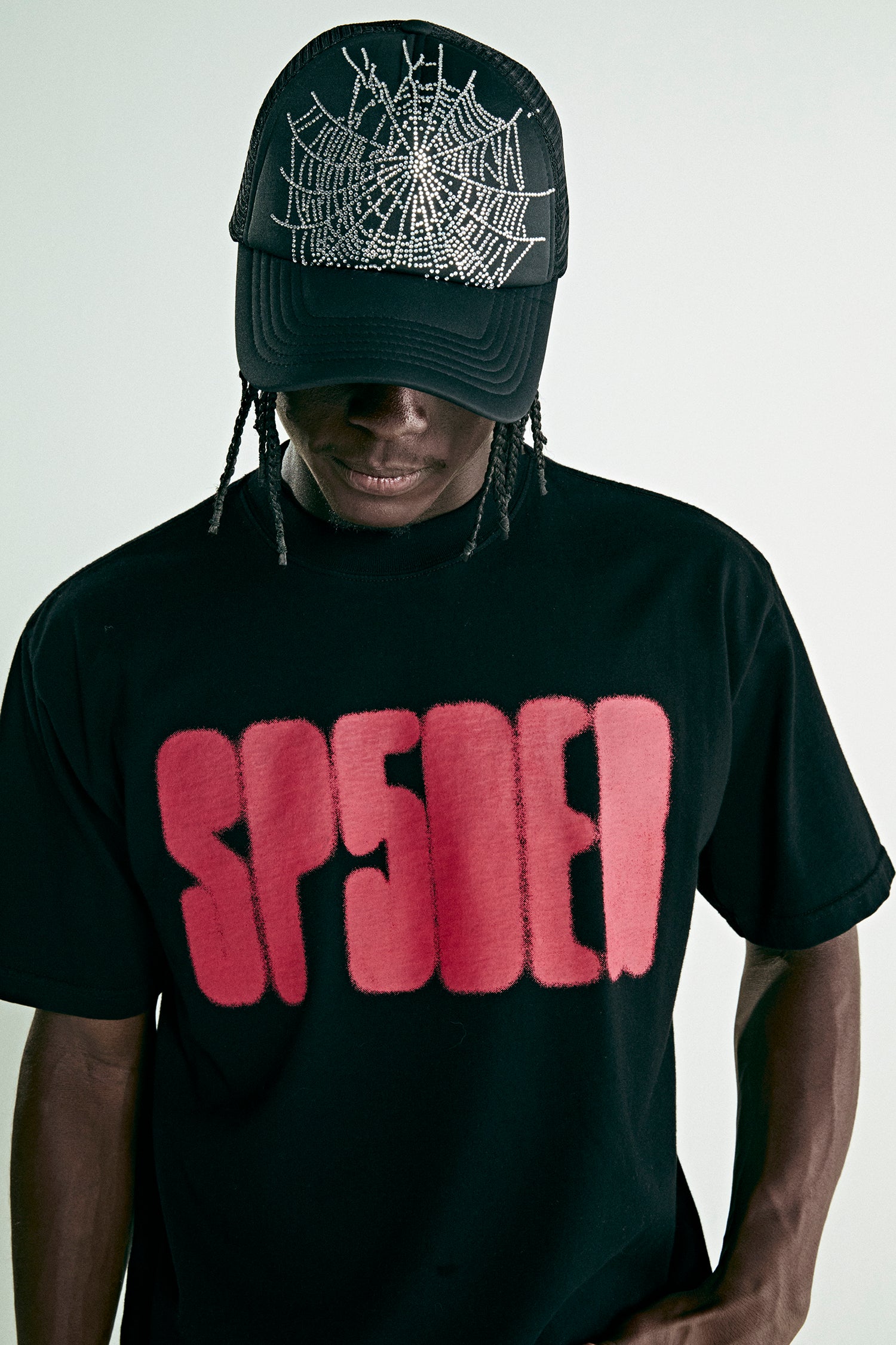 Black cotton tee with red 'SP5DER' text print, worn by a person in a black rhinestone spiderweb trucker hat.