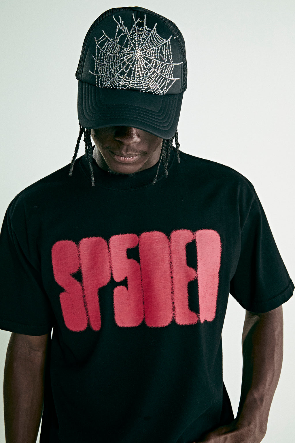 Black cotton tee with red 'SP5DER' text print, worn by a person in a black rhinestone spiderweb trucker hat.