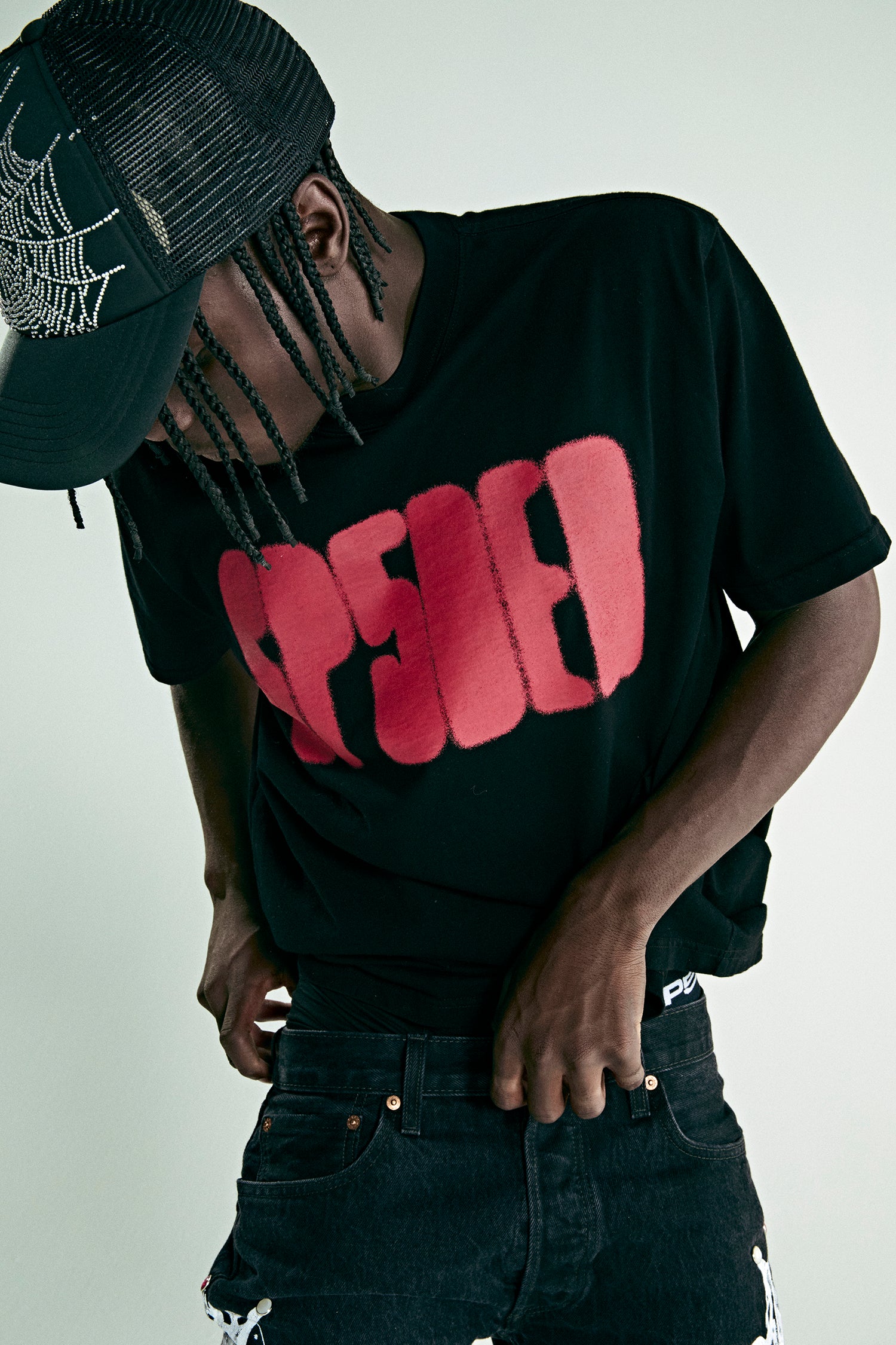 Black cotton tee with red 'SP5DER' text, worn by a person in a spiderweb trucker hat.
