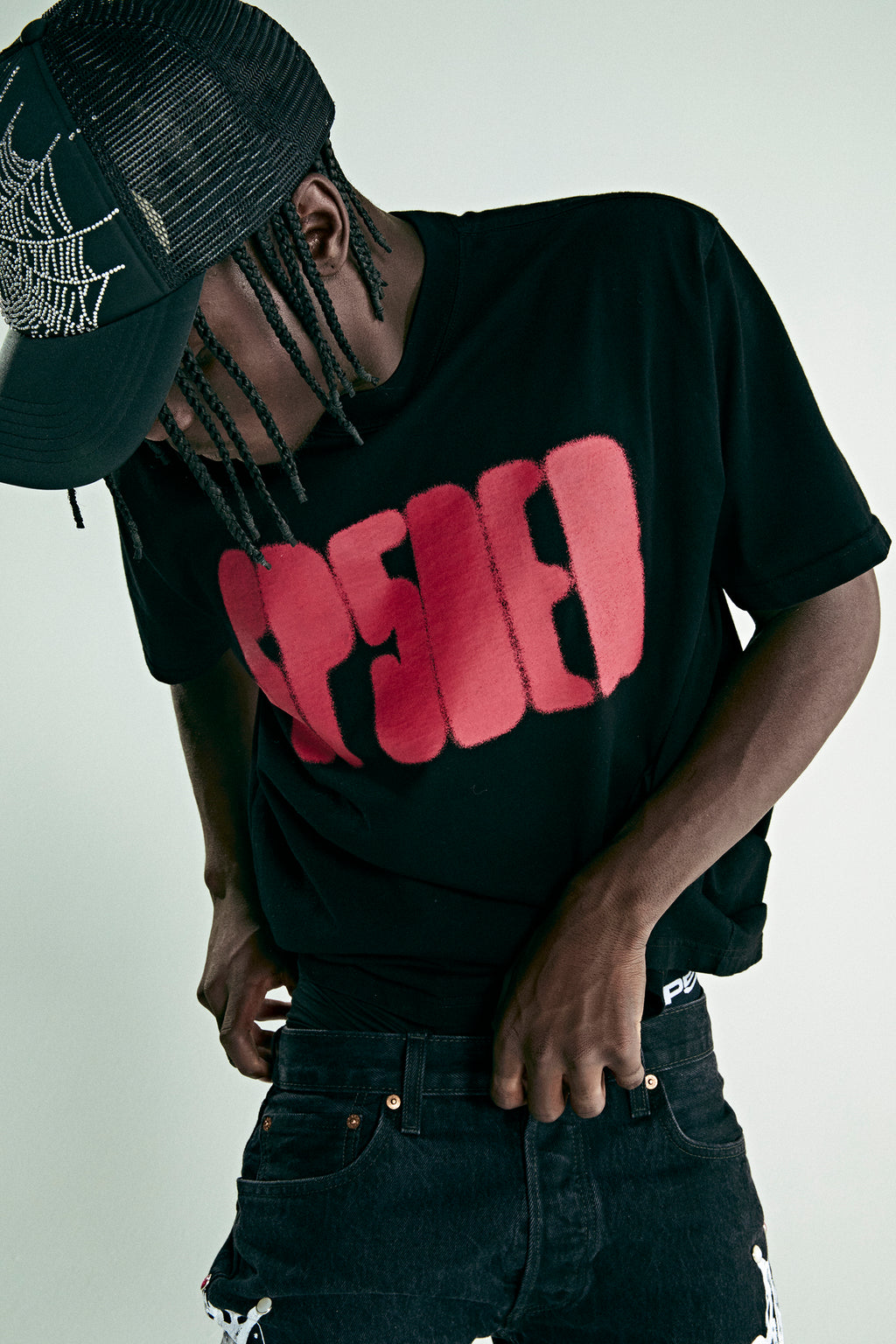 Black cotton tee with red 'SP5DER' text, worn by a person in a spiderweb trucker hat.