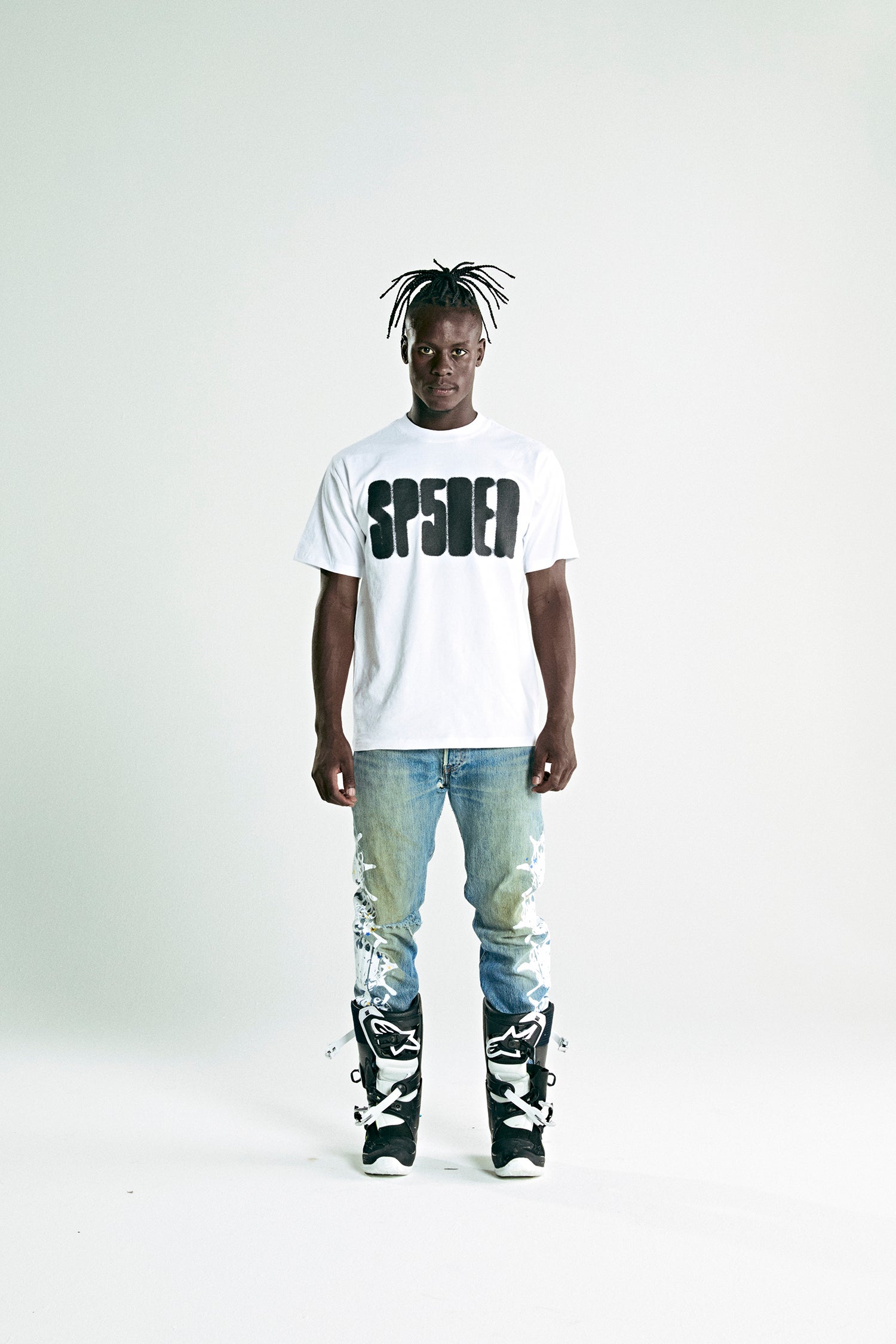 White 100% cotton SP5DER logo tee on a man wearing distressed jeans and black boots.