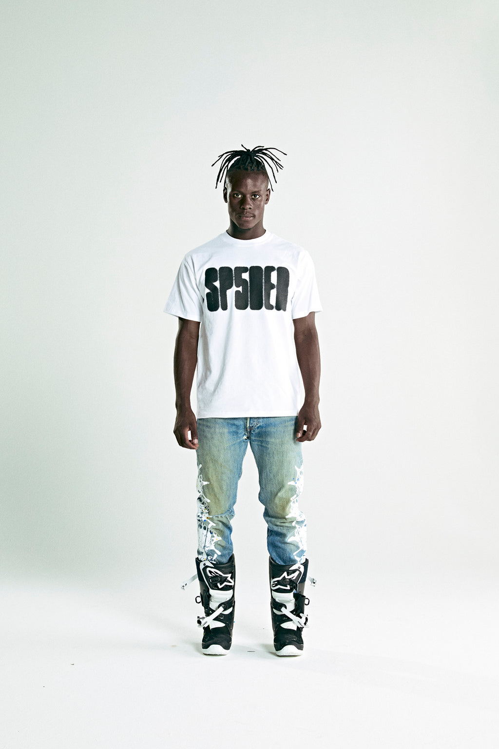 White 100% cotton SP5DER logo tee on a man wearing distressed jeans and black boots.